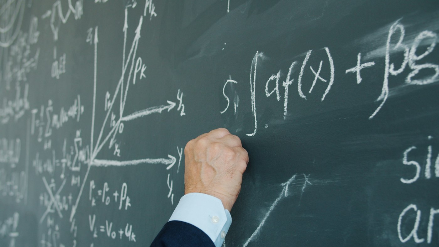 Tangan menulis rumus integral dengan kapur di papan tulis hijau yang penuh notasi matematika.